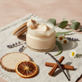 Earth & Ember Foot Butter in open glass jar with wooden lid, light creamy ivory whipped shea butter texture, sandalwood slice, orange slice, vanilla bean, cinnamon sticks, soft neutral linen cloth props